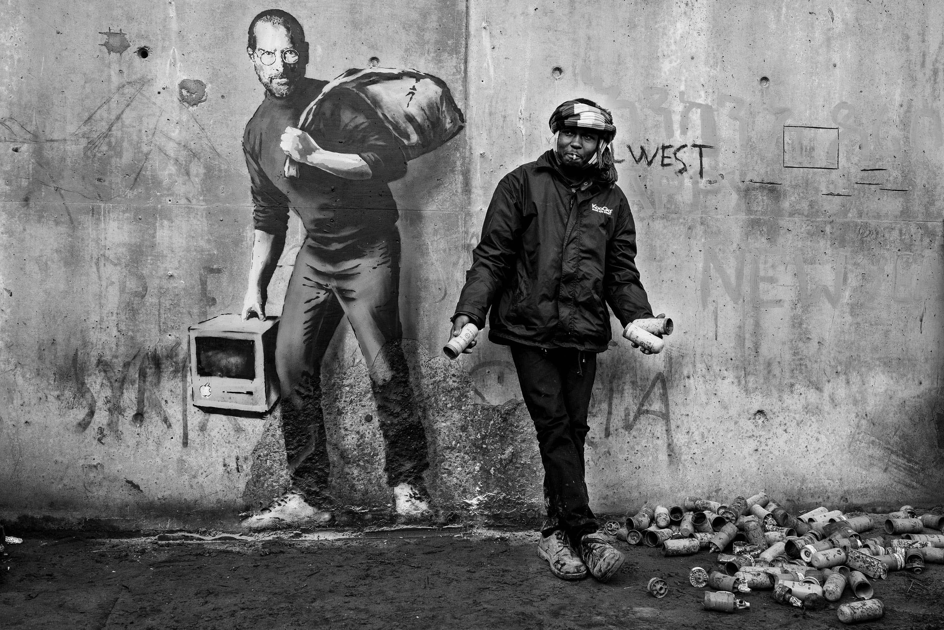 A refugee holds exploded tear gas canisters beside the Banksy mural at the entrance to The Jungle.