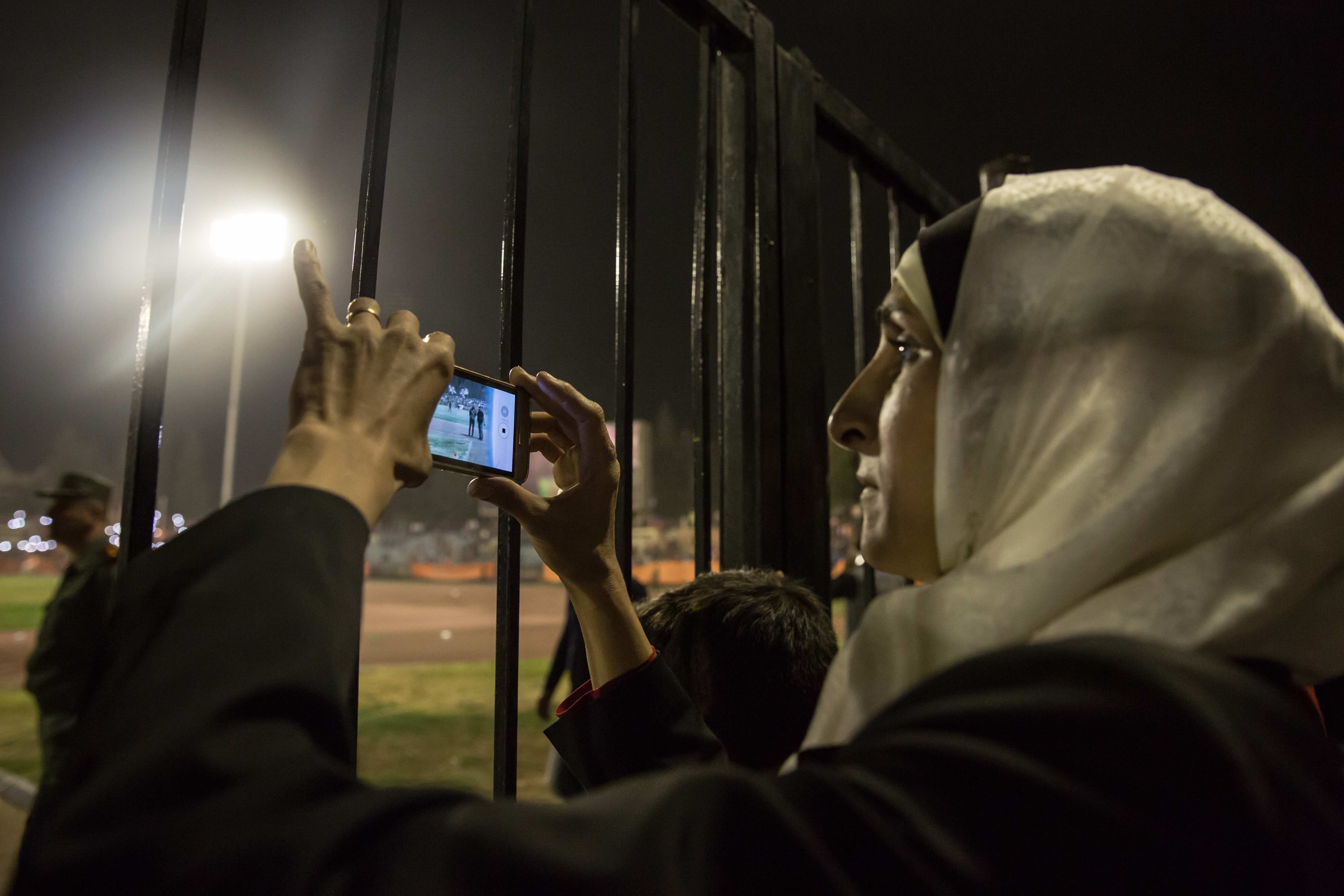 A woman films the pitch. On her phone screen soldiers are more distinctive than football players.